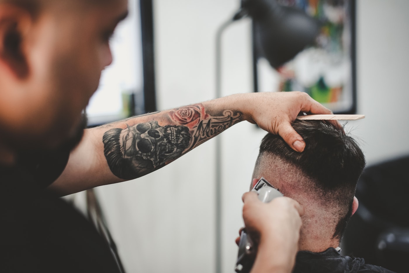 A barber with tattooed arms running clippers over the top of a seated client's head